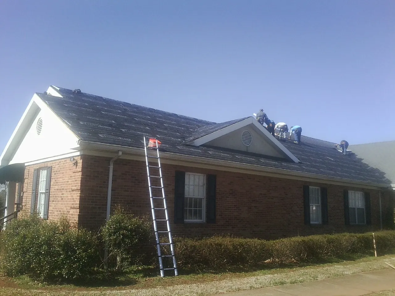 Crew tearing shingles off a brick ranch roof with a ladder set — Emergency Roof Repair work by our crew in West Camp