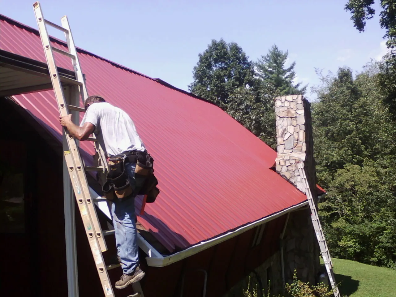 Roofer on a ladder beside a red metal roof and stone chimney — Soffit & Fascia Repair by Roofing Brothers in West Camp